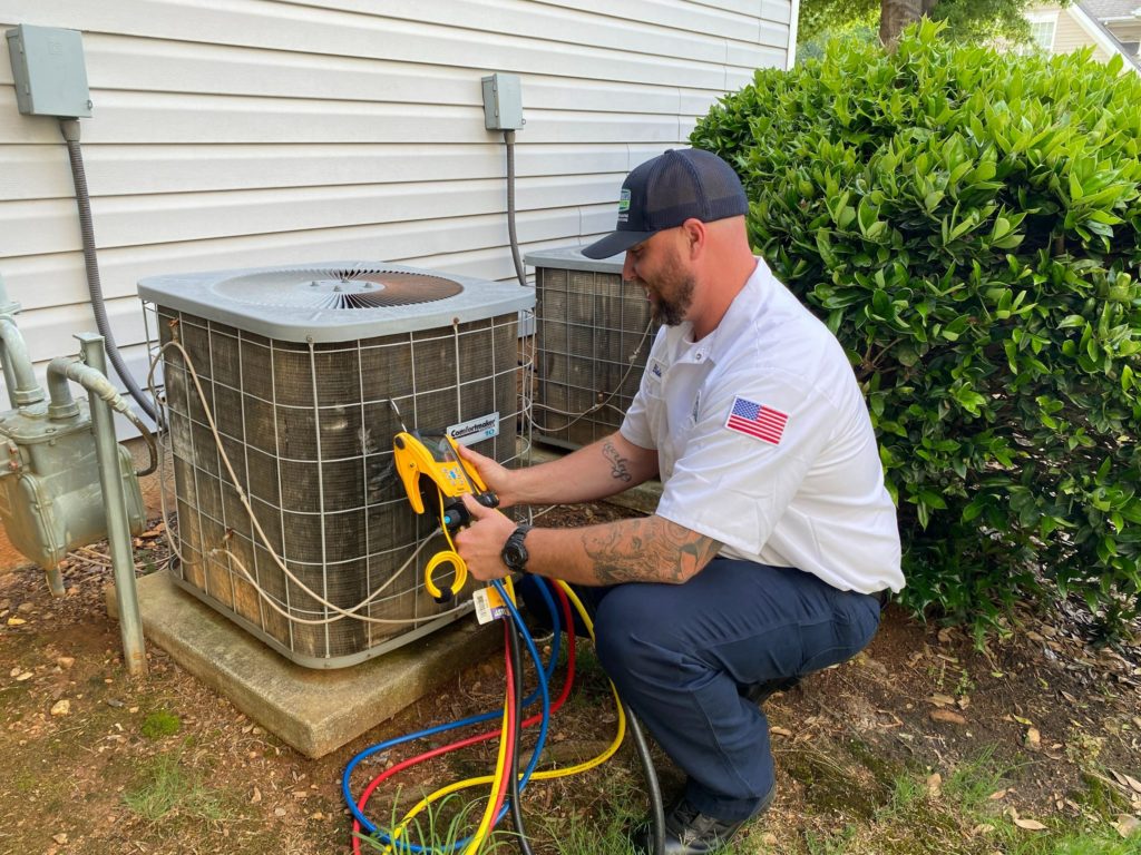 A technician kneels beside an outdoor air conditioning unit, using a yellow tool to perform maintenance.