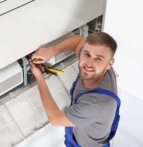 A smiling technician uses pliers to work on the internal components of an air conditioning unit.