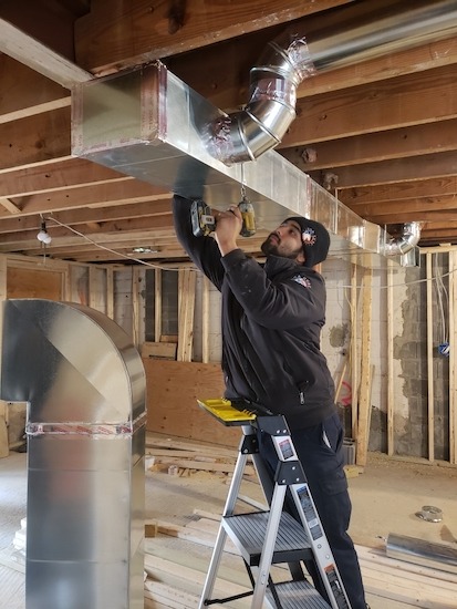 A technician installs ductwork in an unfinished room using a drill.