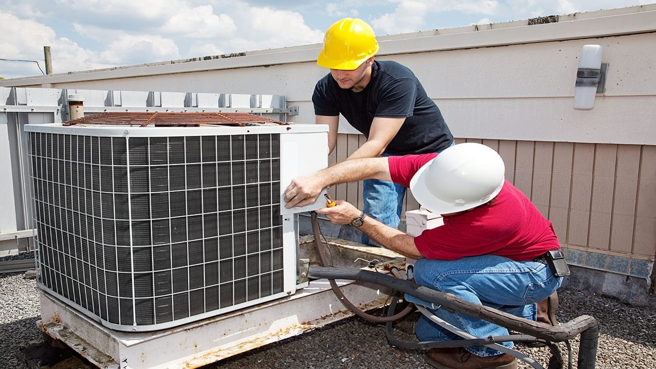 Two workers in hard hats install an outdoor air conditioning unit on a rooftop under a partly cloudy sky.